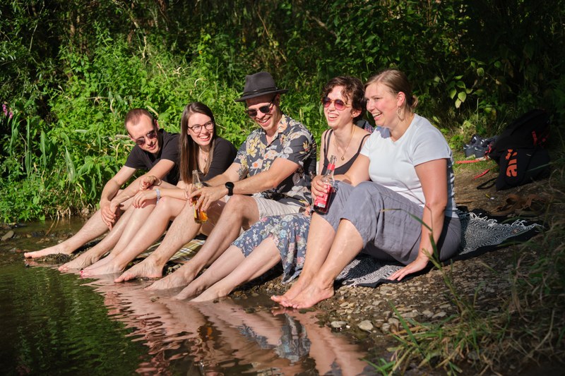 Sechs Studierende sitzen im Sommer an der Lahn und lassen ihre Füße ins Wasser hängen.