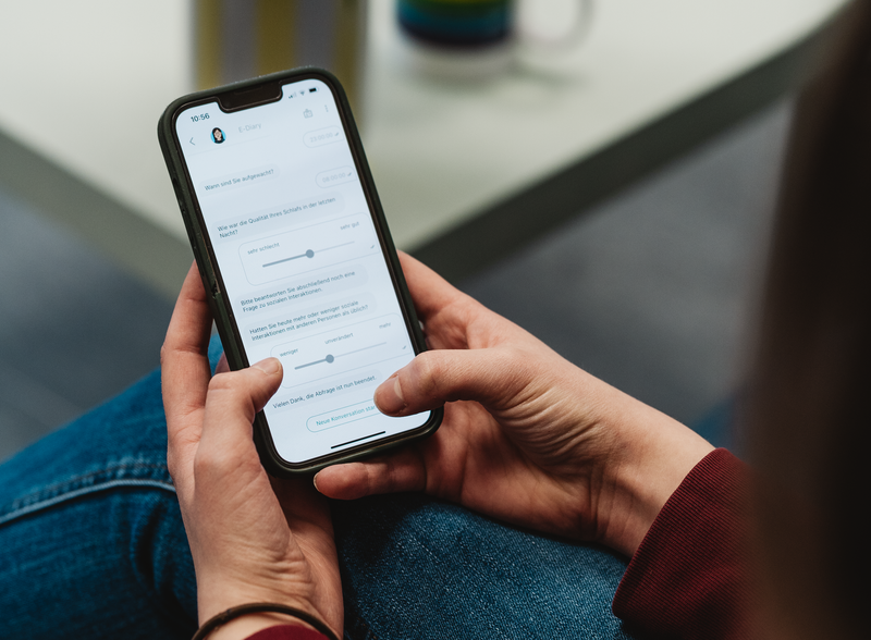 Das Foto zeigt eine Studienteilnehmerin, die mit Hilfe einer EMA(Ecological Momentary Assessment)-App auf einem Smartphone Fragen beantwortet, die sich auf ihren aktuellen Zustand beziehen.