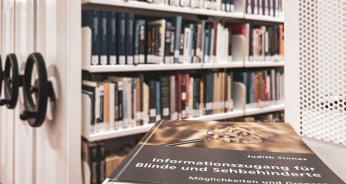 Das Buch "Informationszugang für Blinde- und Sehbehinderte" von Judith Tinnes vor dem Kompaktusregal im Untergeschoss der Universitätsbibliothek