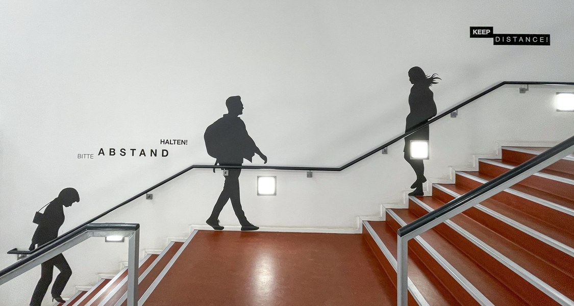 Aufgeklebte Silhouetten, die zum Abstand halten auffordern an der Wand hinter der Haupttreppe der UB