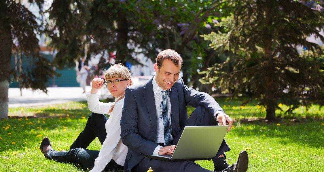 Mann mit Laptop auf dem Schoß und Frau sitzen nebeneinander im Garten und blicken auf den Bildschirm.