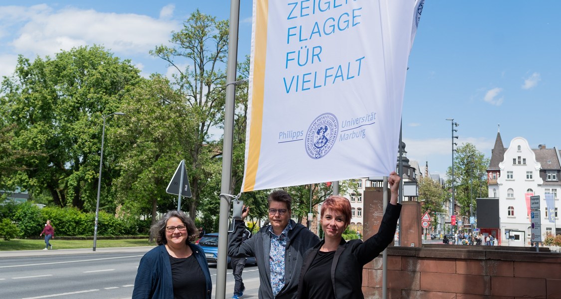 Drei Personen stehen unter einem Fahnenmast an dem die Flagge zum Deutschen Diversity Tag gehisst wurde.