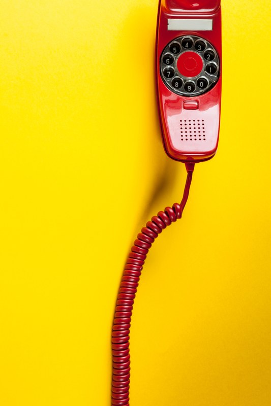 Gelber Hintergrund, darauf ein roter Telefonhörer. Der Hörer hat ein rotes Kabel das nach unten hängt. Auf der Innenseite des Hörers ist ein Wahlkreis mit den Zahlen 1-9 und 0.