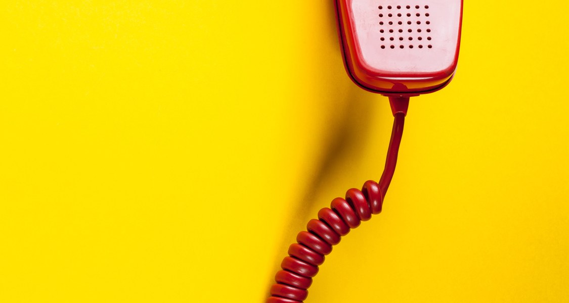 Gelber Hintergrund, darauf ein roter Telefonhörer. Der Hörer hat ein rotes Kabel das nach unten hängt. Auf der Innenseite des Hörers ist ein Wahlkreis mit den Zahlen 1-9 und 0.
