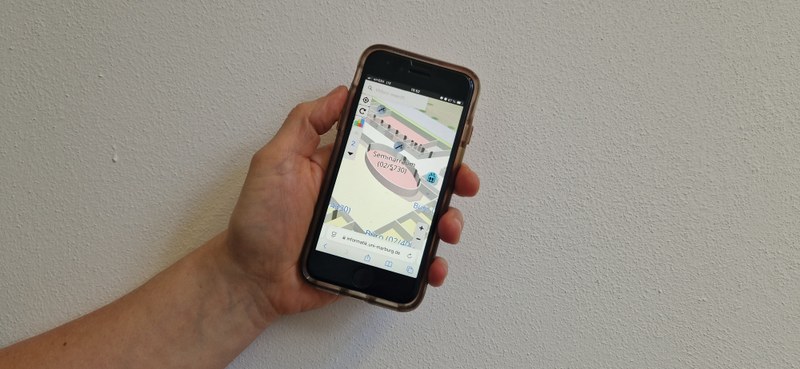 Eine Hand hält ein Smartphone auf dem die Anwendung MINKA einen Seminarraum zeigt