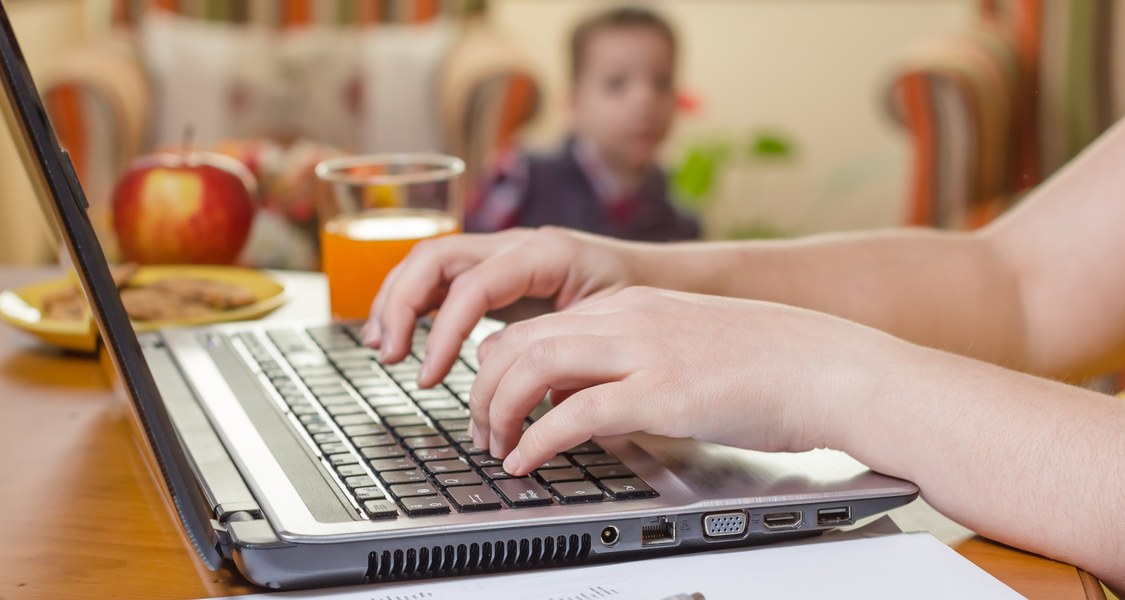 Das Bild zeigt eine Tastatur eines Laptops, auf der gerade eine Person etwas eintippt. Der Laptop steht auf einem Tisch und im Hintergrund ist ein Glas mit Fruchtsaft, ein Apfel und ein Kind zu erkennen.