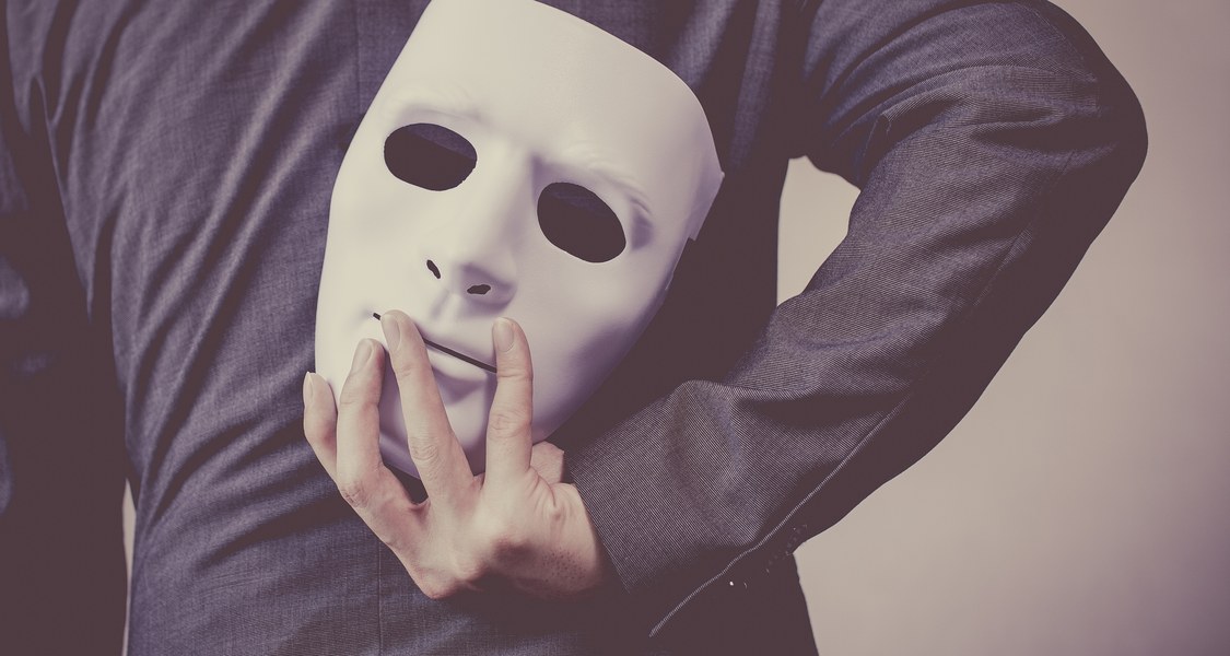 Auf dem Bild ist eine weiße Gesichtsmaske zu sehen, die jemand hinter den Rücken hält.