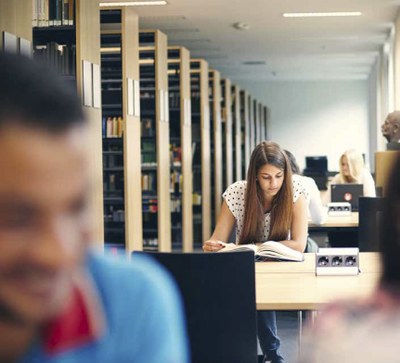 Studierende lesen in Bibliothek. Person im nahen Vordergrund ist unfokussiert, Person weiter hinten ist im Fokus.