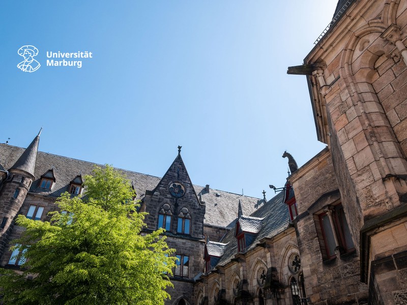 Blick auf Fassade und Dächer der Alten Universität bei strahlend blauem Himmel; oben links das neue Logo der Uni Marburg in Weiß
