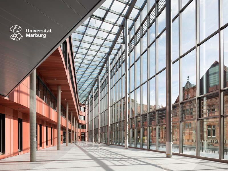 Atrium der Universitätsbibliothek, durch die Glasfront rechts sieht man den historischen Schäferbau; oben links das neue Logo der Uni Marburg in Weiß