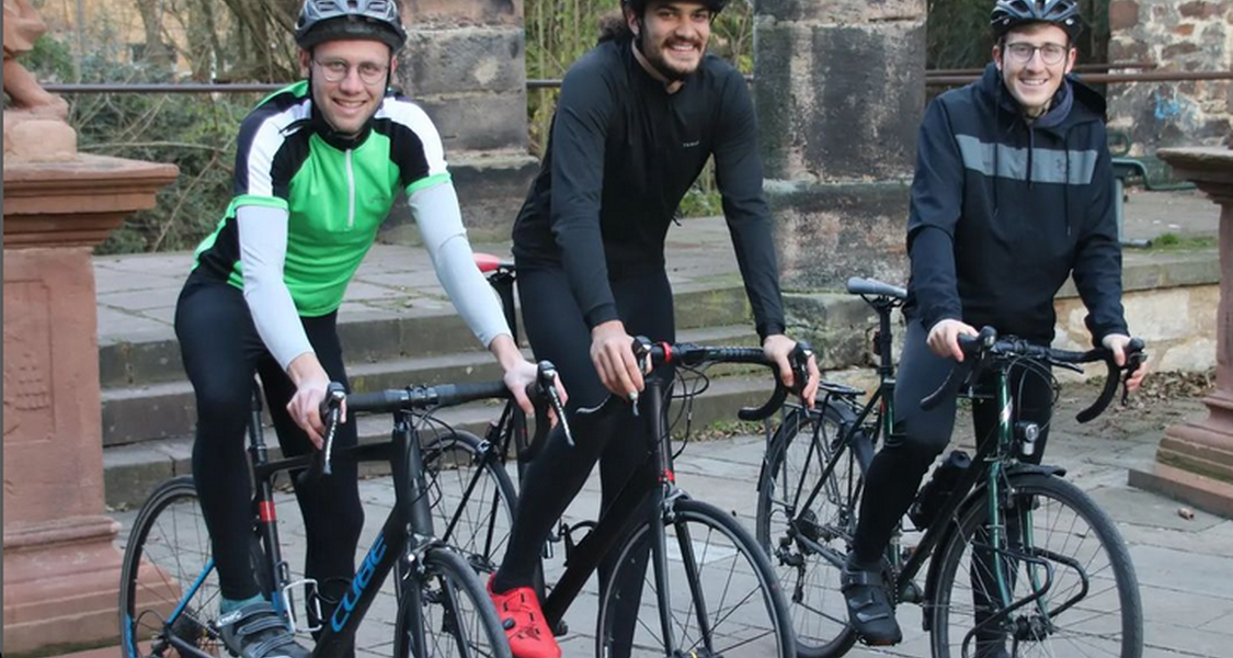 Die drei Studenten auf ihren Fahrrädern stehen vor der Elisabethkirche und lächeln in die Kamera.