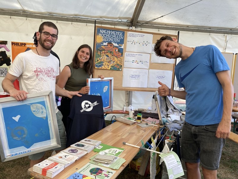 Auf dem Bild sind drei Personen aus dem Green Office abgebildet, die um einen Tisch herumstehen. Auf dem Tisch liegen verschiedene Flyer und Broschüren. Die Personen halten bedruckte T-Shirts und Siebdruckrahmen in den Händen. Im Hintergrund befinden sich Pinnwände mit Plakaten zu nachhaltiger Hochschulgestaltung.