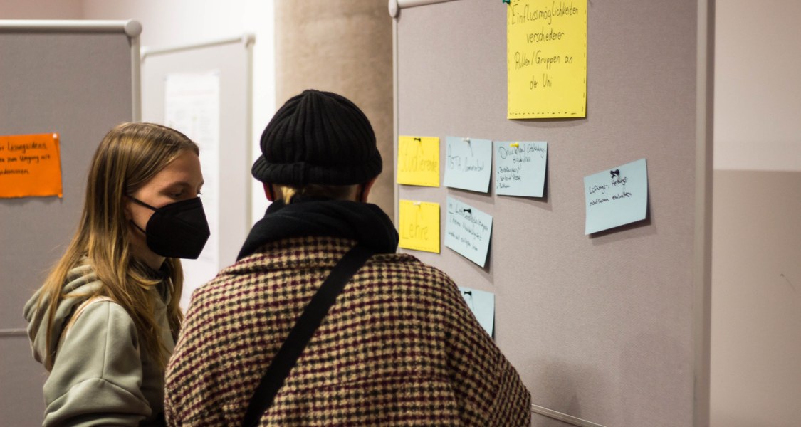 Zwei Personen stehen vor einer Pinnwand und diskutieren angeregt. Auf der Pinnwand steht "Einflussmöglichkeiten verschiedener Gruppen" und Menschen können selbst extra Karten an die Wand hängen, wo sie Einflussmöglichkeiten auf die nachhaltige Entwicklung der Universität sehen.