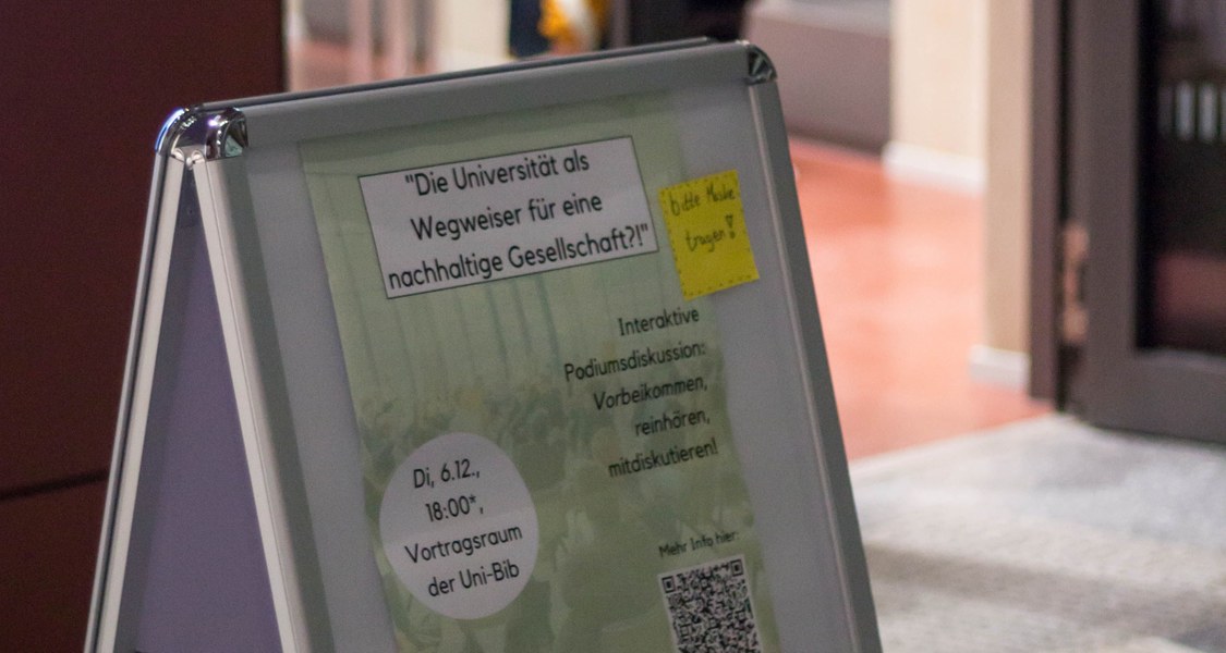 Ein Plakataufsteller steht vor dem Vortragsraum der Unibib. In dem Plakataufsteller hängt ein Poster, was für die interaktive Podiumsdiskussion des Green Offices wirbt.