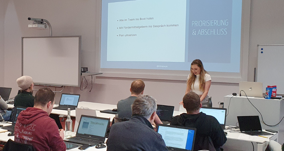Einige Menschen sind mit dem Rücken zu sehen und schauen nach vorne im Schulungsraum der Uni-Bib. Vorne steht die Fördermittelberaterin Mira Pape und erklärt etwas.