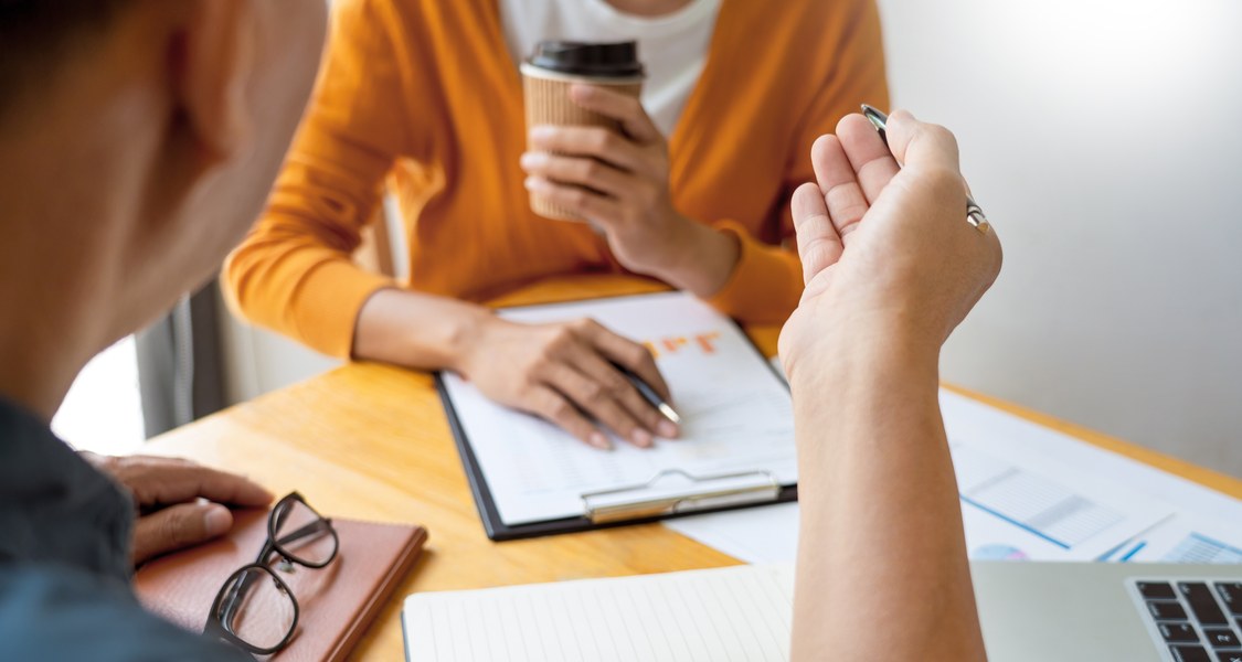 Zwei Personen an einem Besprechungstisch mit aufgeklapptem Laptop, Kaffeebecher, Brille und Notizbüchern