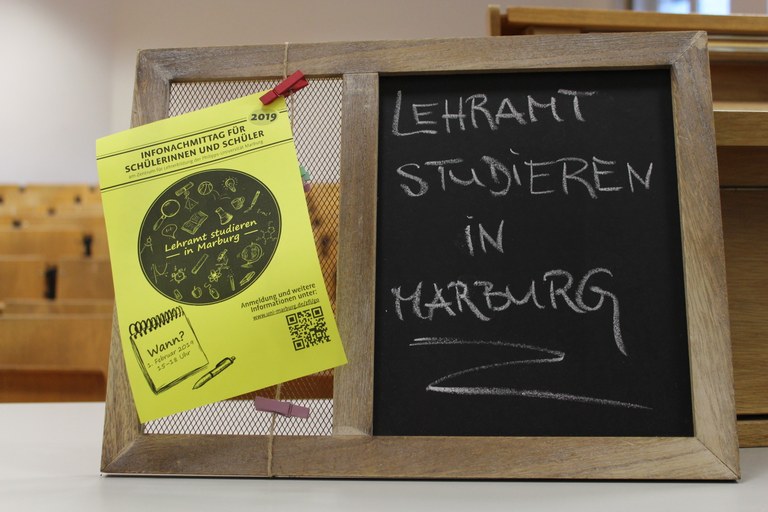 Auf dem Pult eines Hörsaals steht eine kleine Pinnwand mit Kreidetafel. Auf der Tafel steht "Lehramt studieren in Marburg" und an die Pinnwand ist ein Flyer für den "Infonachmittag für Schülerinnen und Schüler 2019" geheftet.
