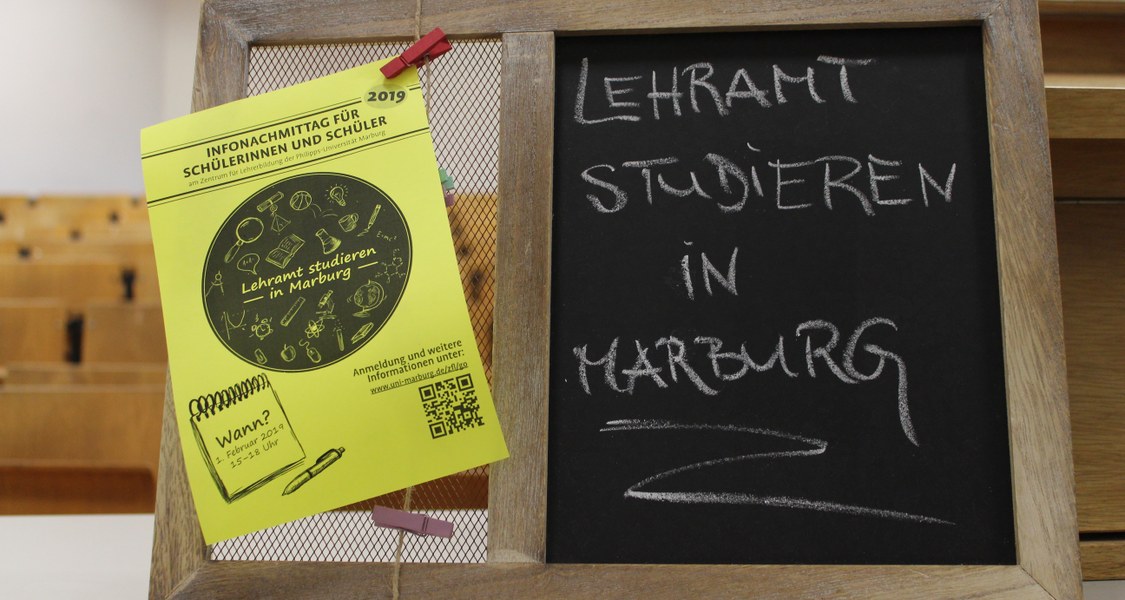Auf dem Pult eines Hörsaals steht eine kleine Pinnwand mit Kreidetafel. Auf der Tafel steht "Lehramt studieren in Marburg" und an die Pinnwand ist ein Flyer für den "Infonachmittag für Schülerinnen und Schüler 2019" geheftet.