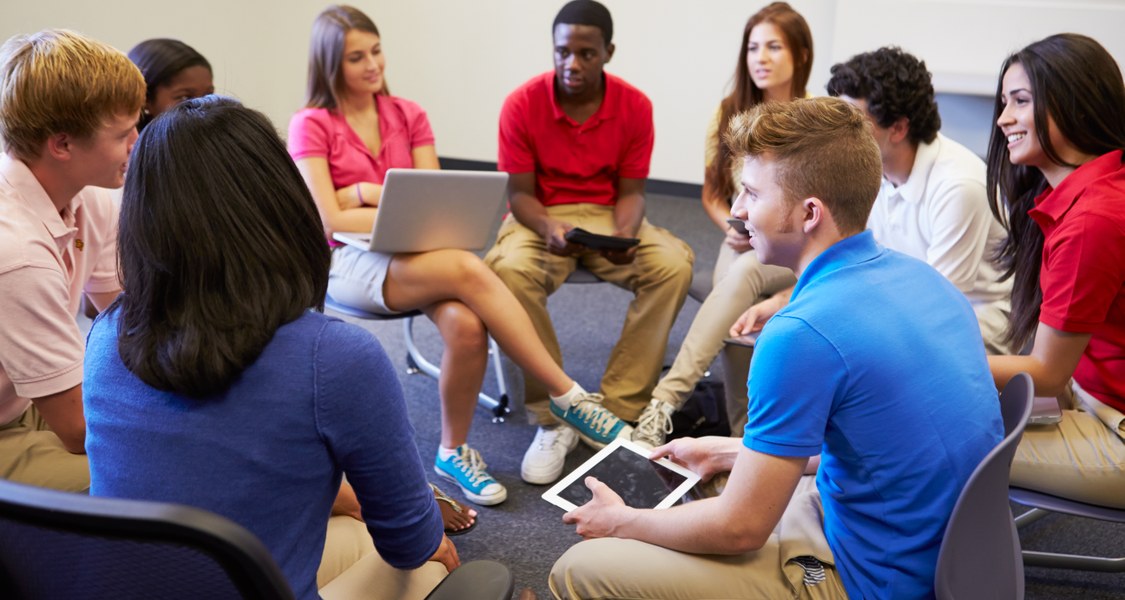 Viele Schüler/innen sitzen in einem Stuhlkreis. Sie halten verschiedene technische Geräte wie Tablets oder Laptops in den Händen.