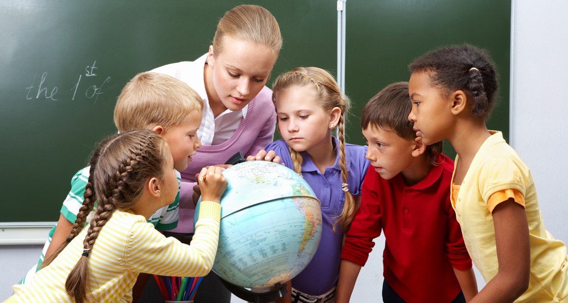 Eine Lehrerin steht mit fünf Kindern um einen Globus auf dem Pult herum und schauen sich diesen an.