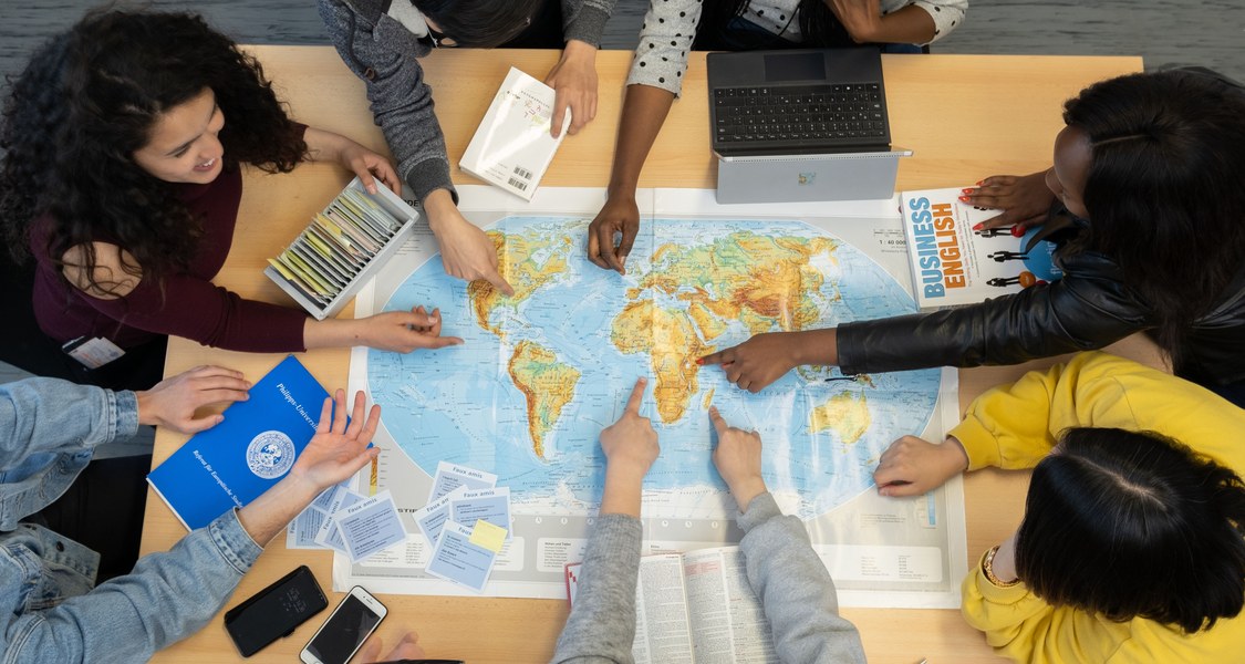 Weltkarte auf einem Tisch, um welche mehrere Studierende herum sitzen und mit dem Finger zeigen.