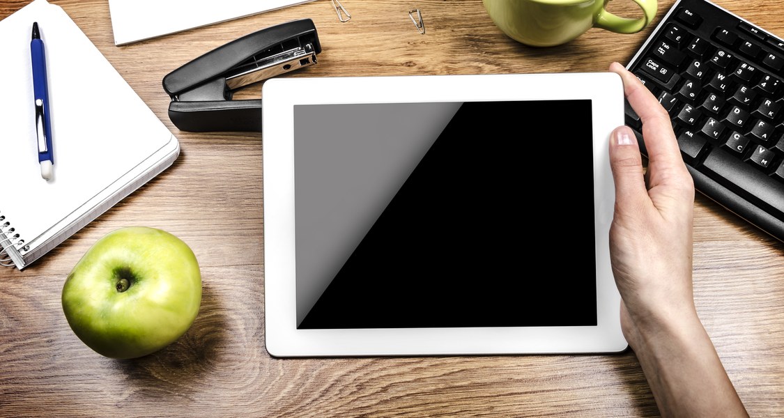 Auf einem Schreibtisch hält eine Person ein Tablet in der Hand; rund herum befinden sich auf dem Schreibtisch ein Apfel, ein Notizblock mit Stift, ein Tacker, Büroklammern, eine Tasse und eine Tastatur.