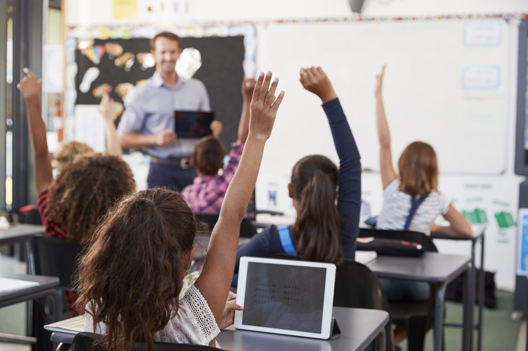 Bild von einer Schulklasse im Klassenzimmer. Im Hintergrund steht die Lehrkraft mit Tablet an der Tafel; der Fokus liegt auf den Schülerinnen und Schülern, welche ebenfalls Tablets bedienen und sich melden.