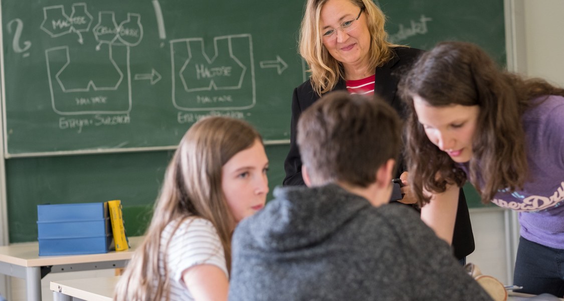 Gruppenarbeit von drei Schülerinnen und Schülern im Chemie-Klassenraum, welche von einer Lehrkraft beobachtet wird.