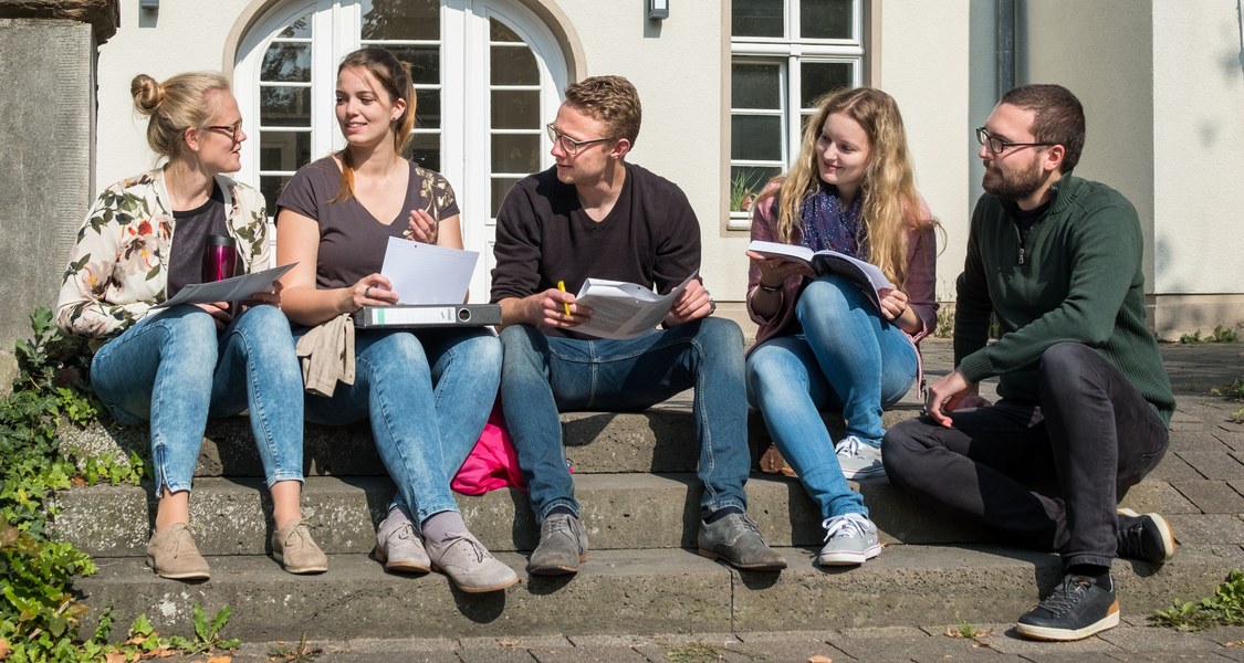 Fünf Studierende sitzen als Arbeitsgruppe mit Arbeitsblättern, Büchern, Stiften, Kaffeebecher und Ordner draußen vor einem Universitätsgebäude.