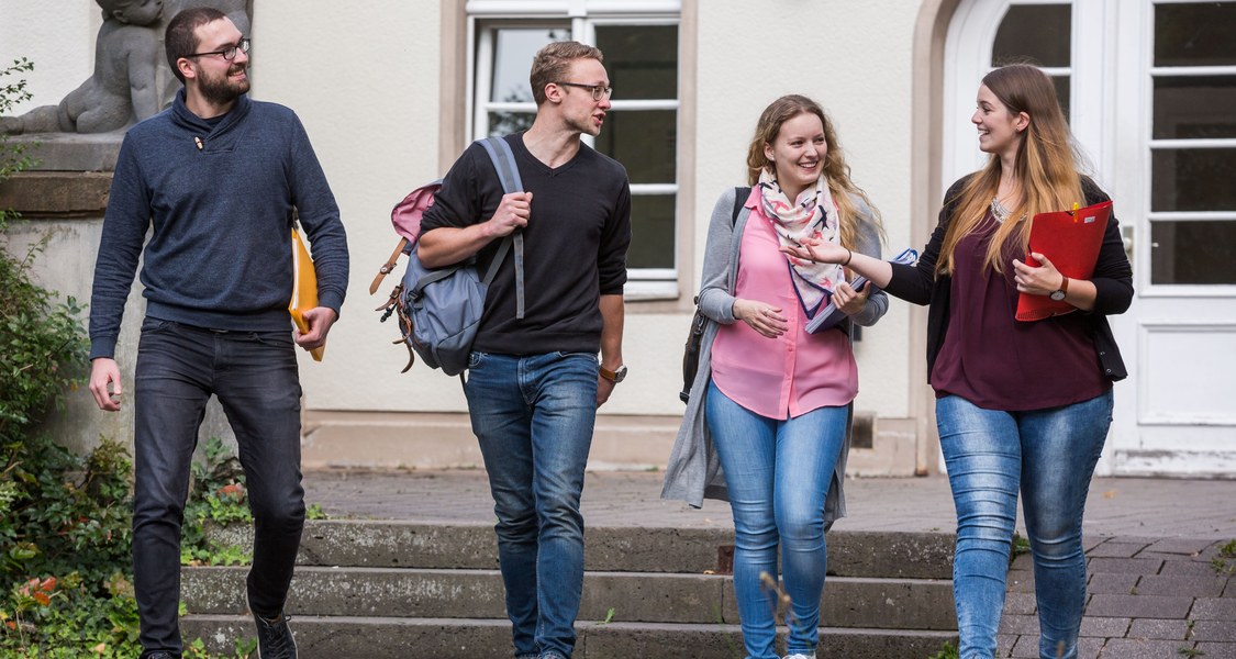 Vier Studierende draußen vor dem Carolinenhaus mit Rucksäcken und Ordnern in der Hand gehend und sich unterhaltend.