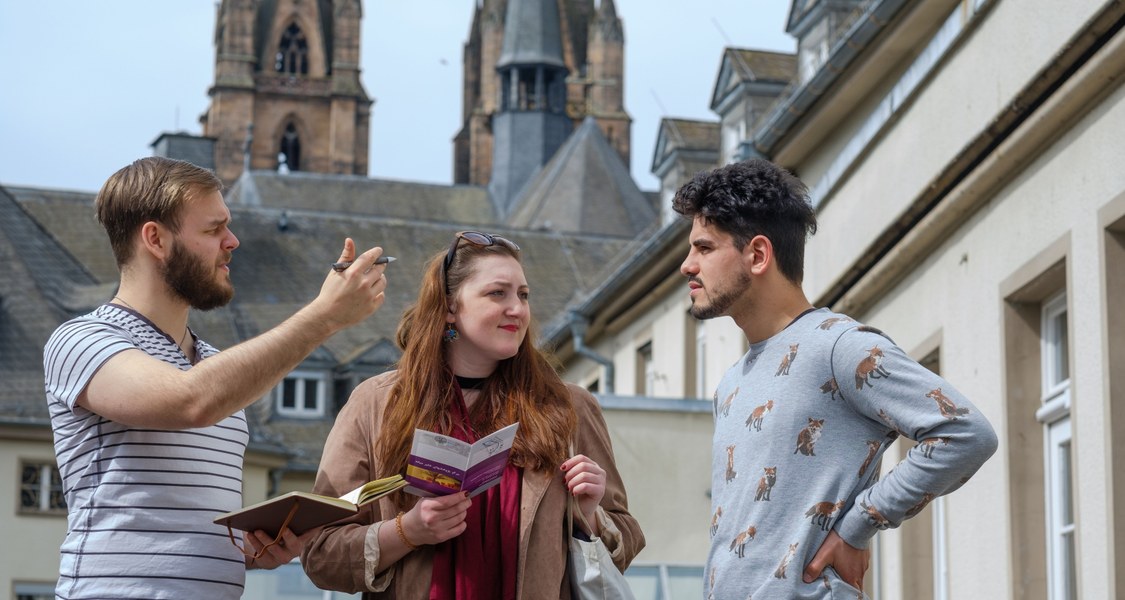 Drei Studierende mit diversen kulturellen Hintergründen stehen im Innenhof der Deutschhausstraße, einer gestikuliert. Im Hintergrund sieht man die Elisabethkirche.