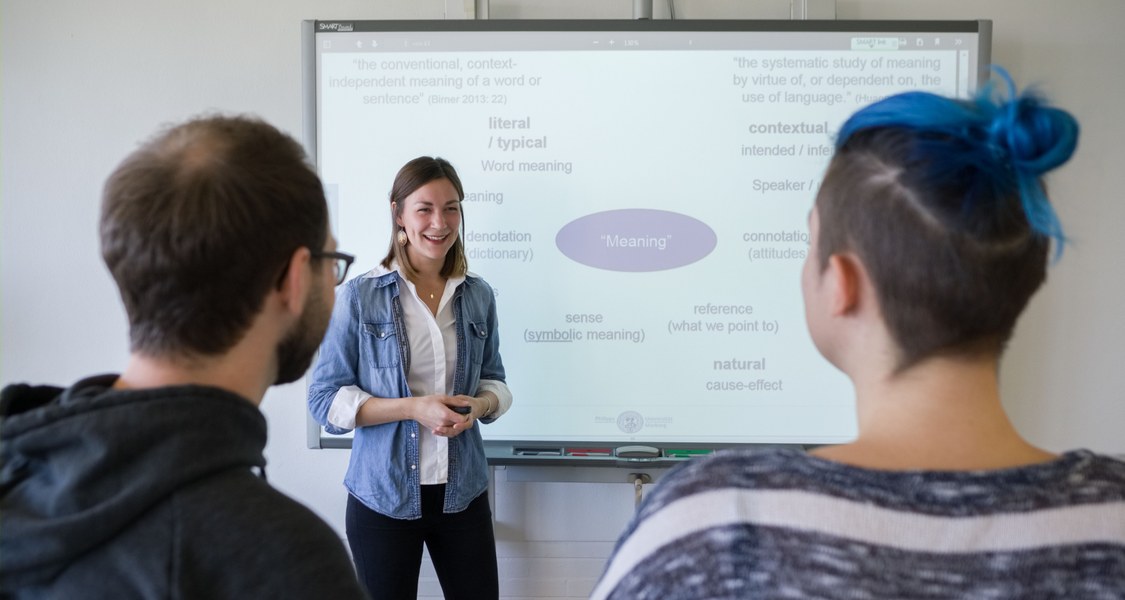 Eine weiblich gelesene Person steht vor einem Smartboard, im Vordergrund sind zwei Personen von hinten zu sehen, die ihr zuhören.