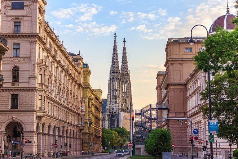 Man blickt einen Abschnitt der Ringstraße hinunter. Rechts und links der Straße befinden sich fünfstöckige hellbraune Gebäude im Stil der Neorenaissance und am Ende der Straße, im Zentrum des Bildes befindet sich die im Stil der Neogotik gebaute Votivkirche. Der Himmel hinter der Kirche ist noch vom Sonnenuntergang orange gefärbt.