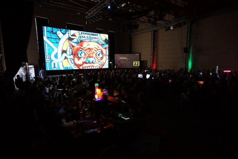 A darkened hall with many people at tables using computers. In the back of the hall is a huge canvas showing a Demo.