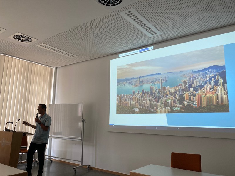 Bild eines Vortrags. Im Hintergrund sieht man Folien mit dem Bild HongKongs an die Wand projiziert.