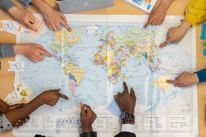 Depicted is a map of the world with several hands of different skin-coloured people pointing to different regions on the map