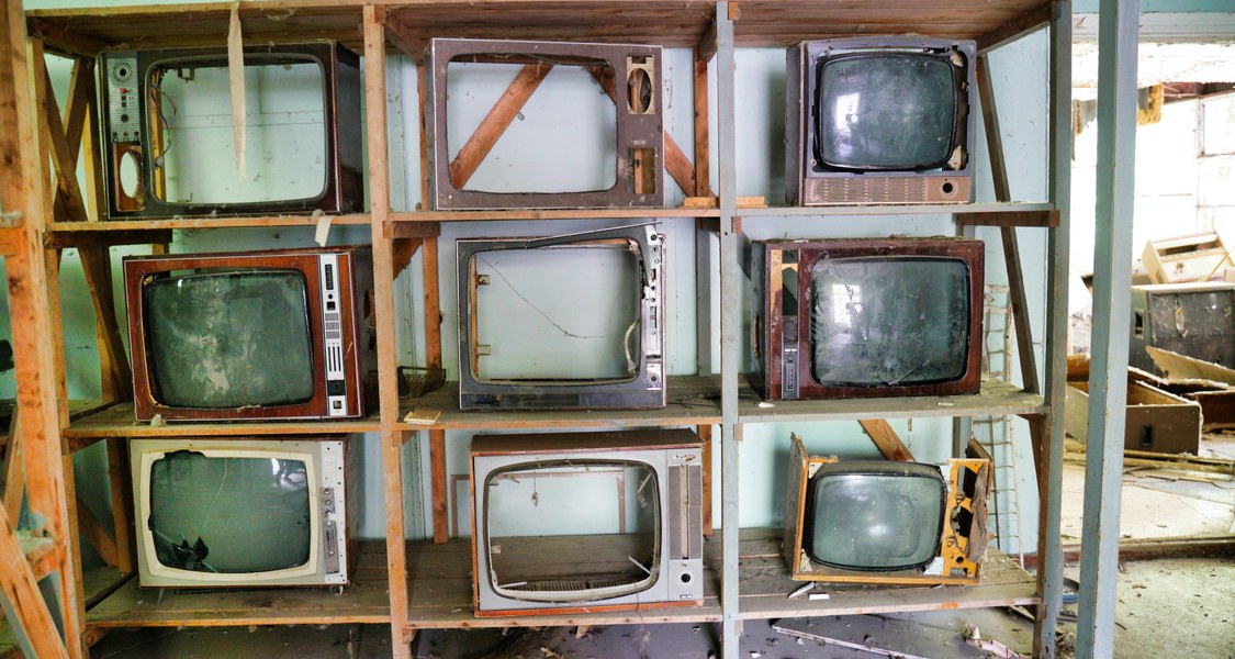 A dilapidated room with an shelf containing 9 broken televisions.