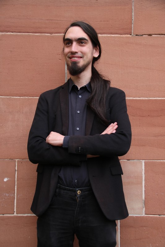 A full-length portrait of Adrian Kinzig standing in front of a brick wall