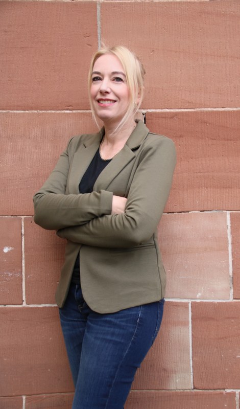 A full-length portrait of Sabine Neumann standing in front of a brick wall