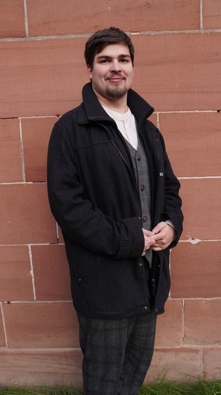 A full-length portrait of Tom Wirtz standing in front of a brick wall