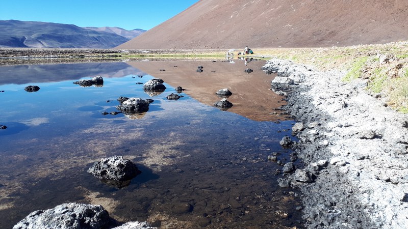 Flat mat and microbialites in a lake. Inactive volcanoes are in the background. Scientists set up experiments and a camp at the coast.