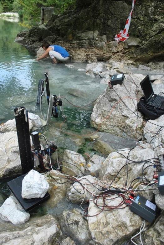 Scientist working in a river. In the forefront, there are microsensor setups for measurements in green biofilms connected to analytical tools via many cables. The scientist in the background is collecting water samples.