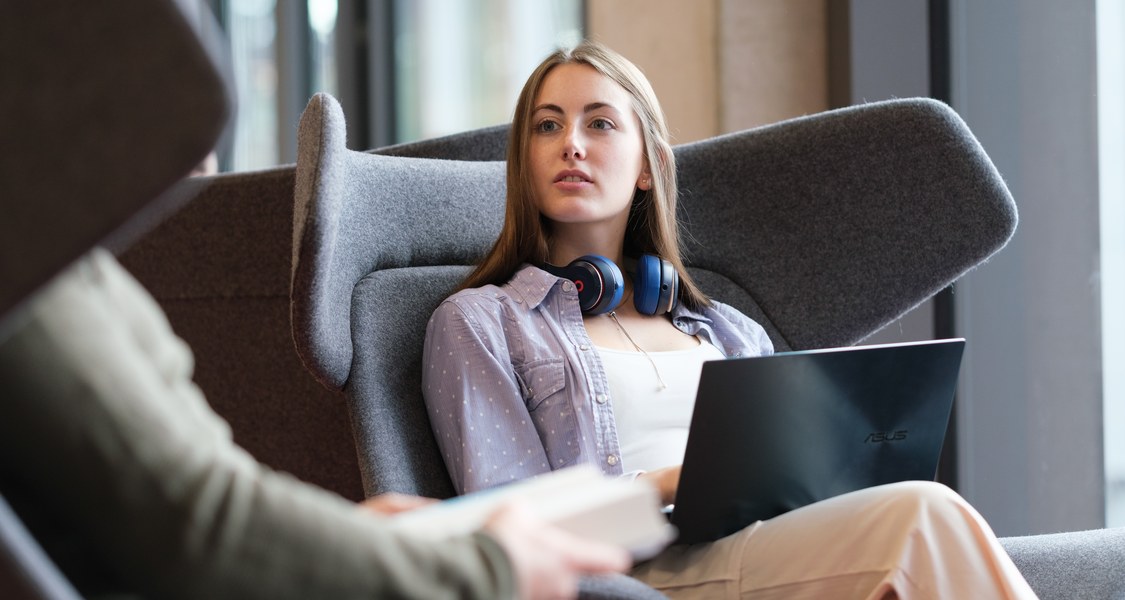 A female student is sitting with her laptop in a high grey armchair. She is having a conversation with a person sitting across from her.
