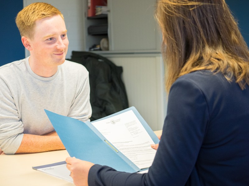 A study advisor and a person seeking advice sit across from each other, discussing a document with the university seal visible.