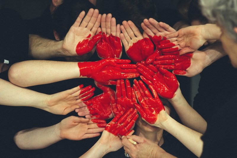 One large red heart is painted across the palms of different people’s hands.