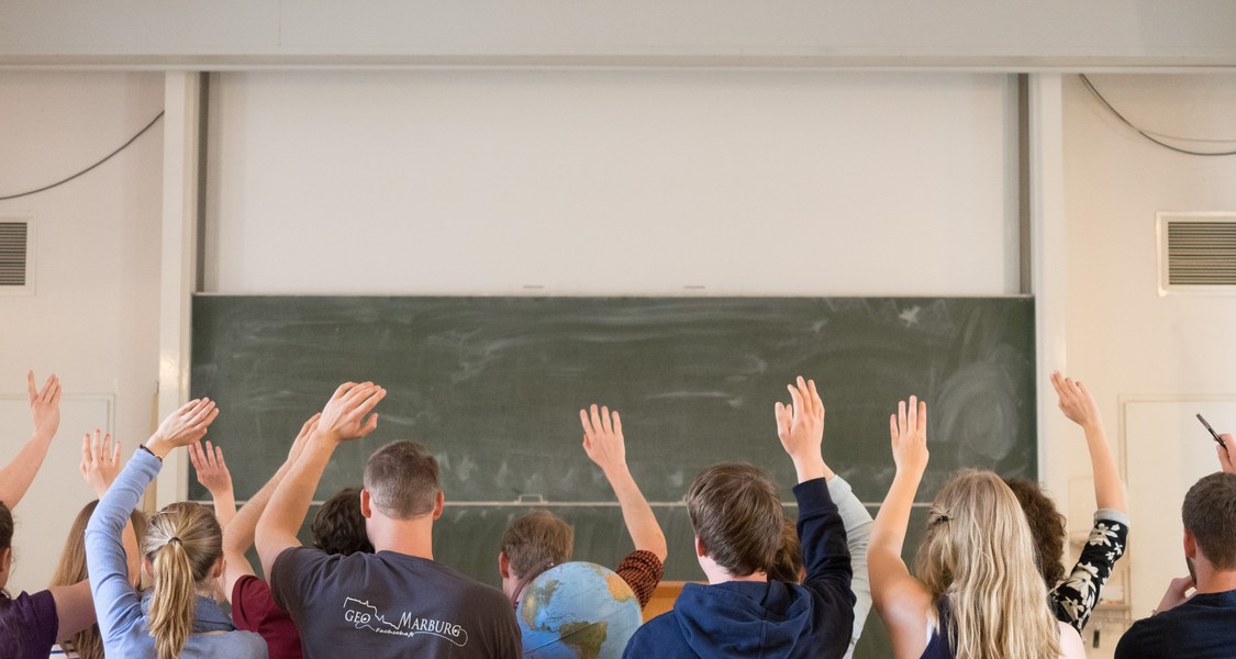 A class of students seated in a lecture hall, raising their hands, with a globe in their midst.