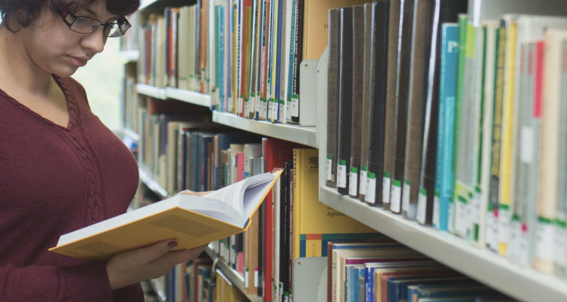 A student standing between bookshelves reads a book.